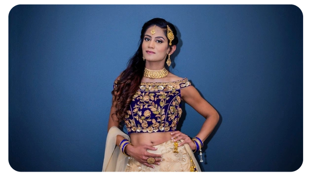 A person poses in a traditional outfit featuring a deep blue, intricately embroidered blouse and a flowing cream skirt against a solid blue background.
