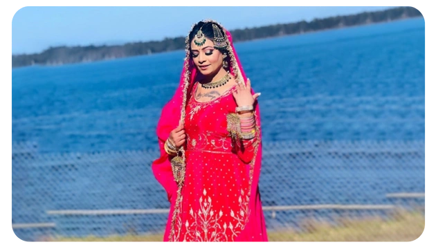 A woman dressed in a vibrant pink traditional outfit, adorned with intricate embroidery, poses by a serene blue waterfront.