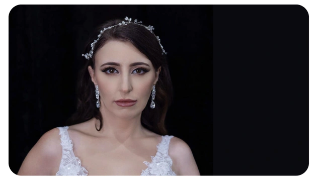 A woman wears a delicate, floral headpiece and an elegant white dress with lace detailing, set against a dark background.