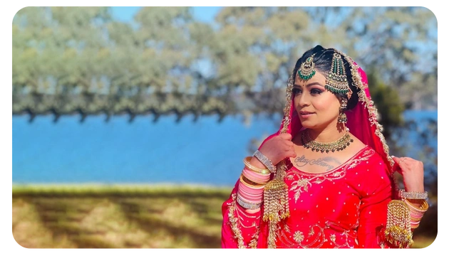 A woman in a vibrant red traditional outfit adorned with intricate jewelry poses gracefully outdoors, with a serene backdrop of trees and water.