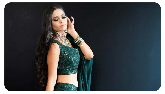 A woman wearing a sparkling green traditional outfit poses elegantly, adorned with jewelry and long, flowing hair against a dark background.