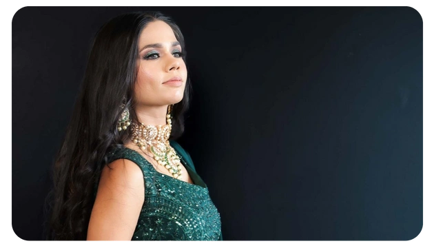 A woman in an elegant green outfit adorned with intricate beadwork and layered pearl jewelry stands against a dark background.