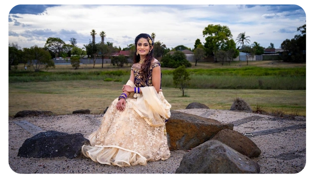 A person in an elegant beige and gold gown sits on rocks, surrounded by a grassy landscape under a cloudy sky.
