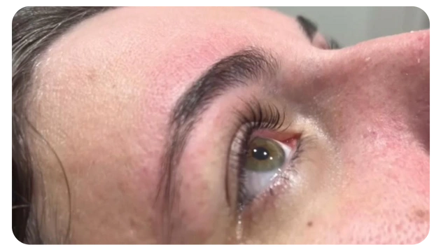 Close-up of a person's eye showcasing long eyelashes, green iris, and well-groomed eyebrow, with slight redness on the skin around.
