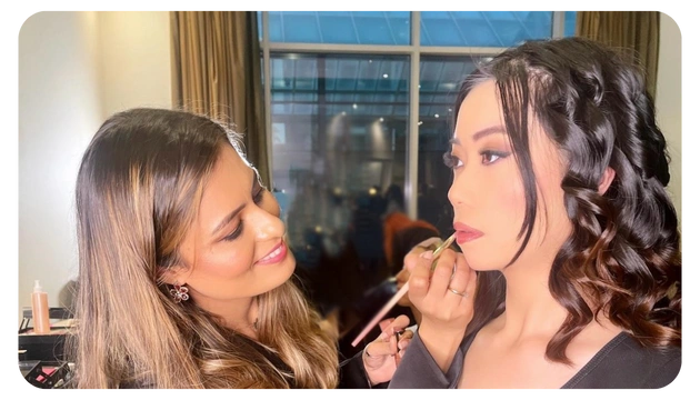 A hairstylist applies makeup to a model in a well-lit room with large windows, showcasing elegant curls and a polished look.