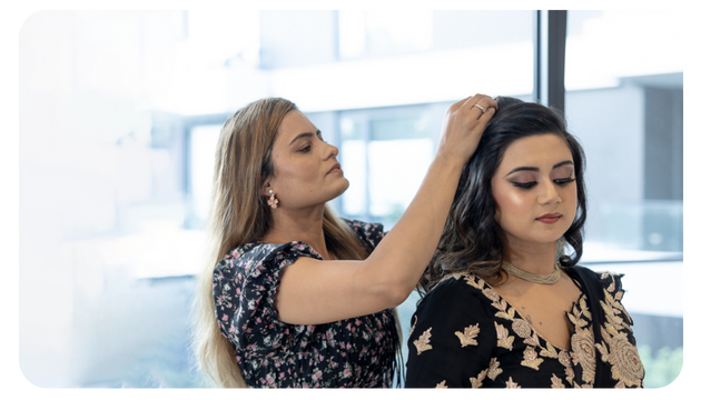 A woman adjusts the hair of another woman in a bright indoor setting, both dressed in elegant attire, suggesting a styling session.