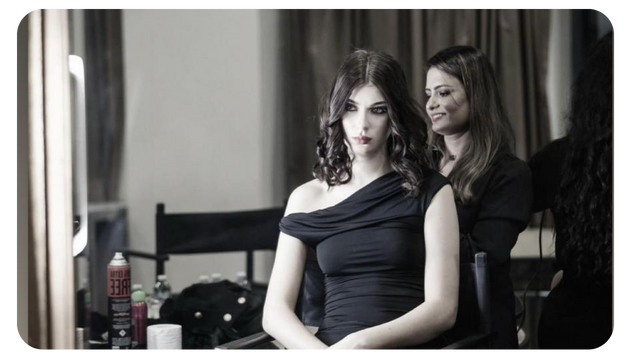 A woman in a black dress sits in a salon chair, while another stylist attends to her hair in a softly lit room.